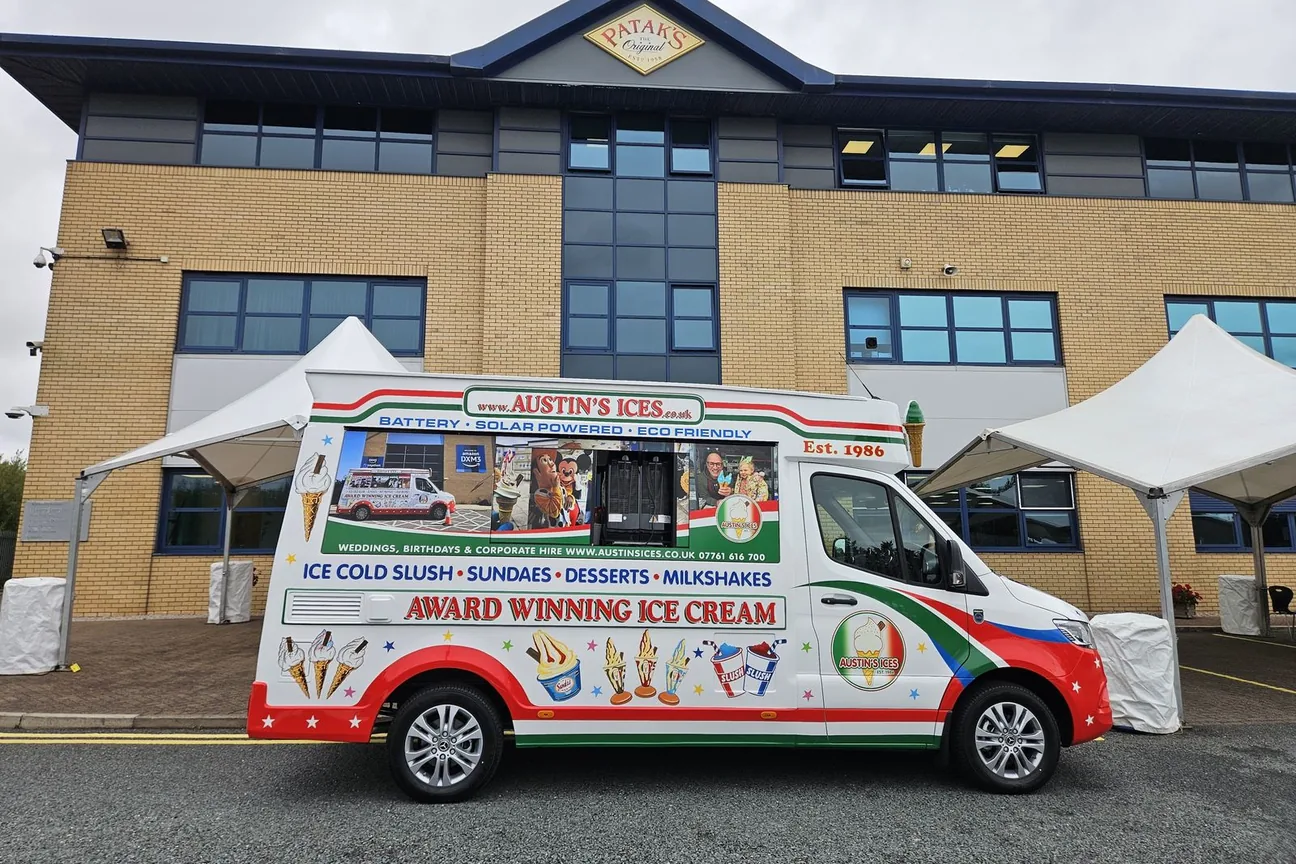 Austins Ices electric ice cream van in profile outside an office building, between two white event gazebos, with the full menu lettering visible